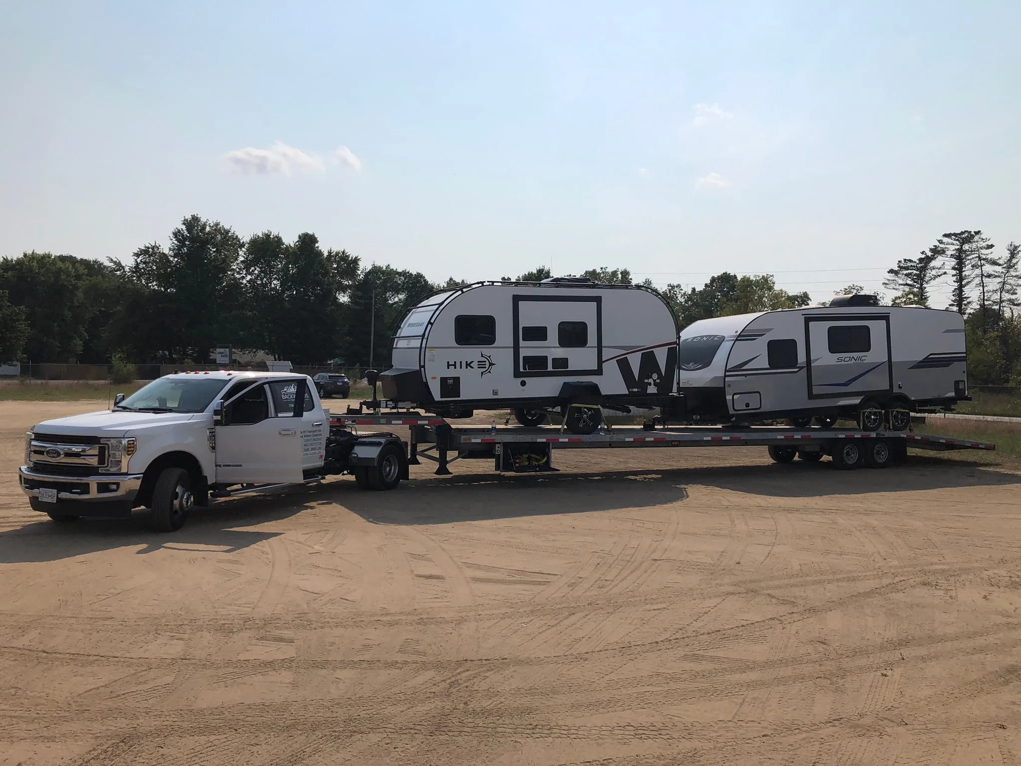 Backwoods pickup with two travel trailers on its deck in a gravel lot